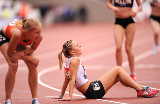 Runner sitting exhausted on a track, another leans forward, both in sports attire. Background shows blurred runners. Mood: tired and intense.
