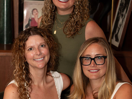 Three women smile warmly in front of a bookcase filled with books and decorative items, complemented by vibrant flowers and personal photographs, two are seated and one is standing.