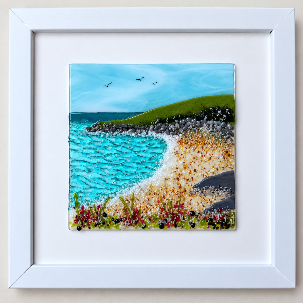 Framed fused glass coastal seascape with turquoise sea, sandy cove, rocky shoreline and green headland.