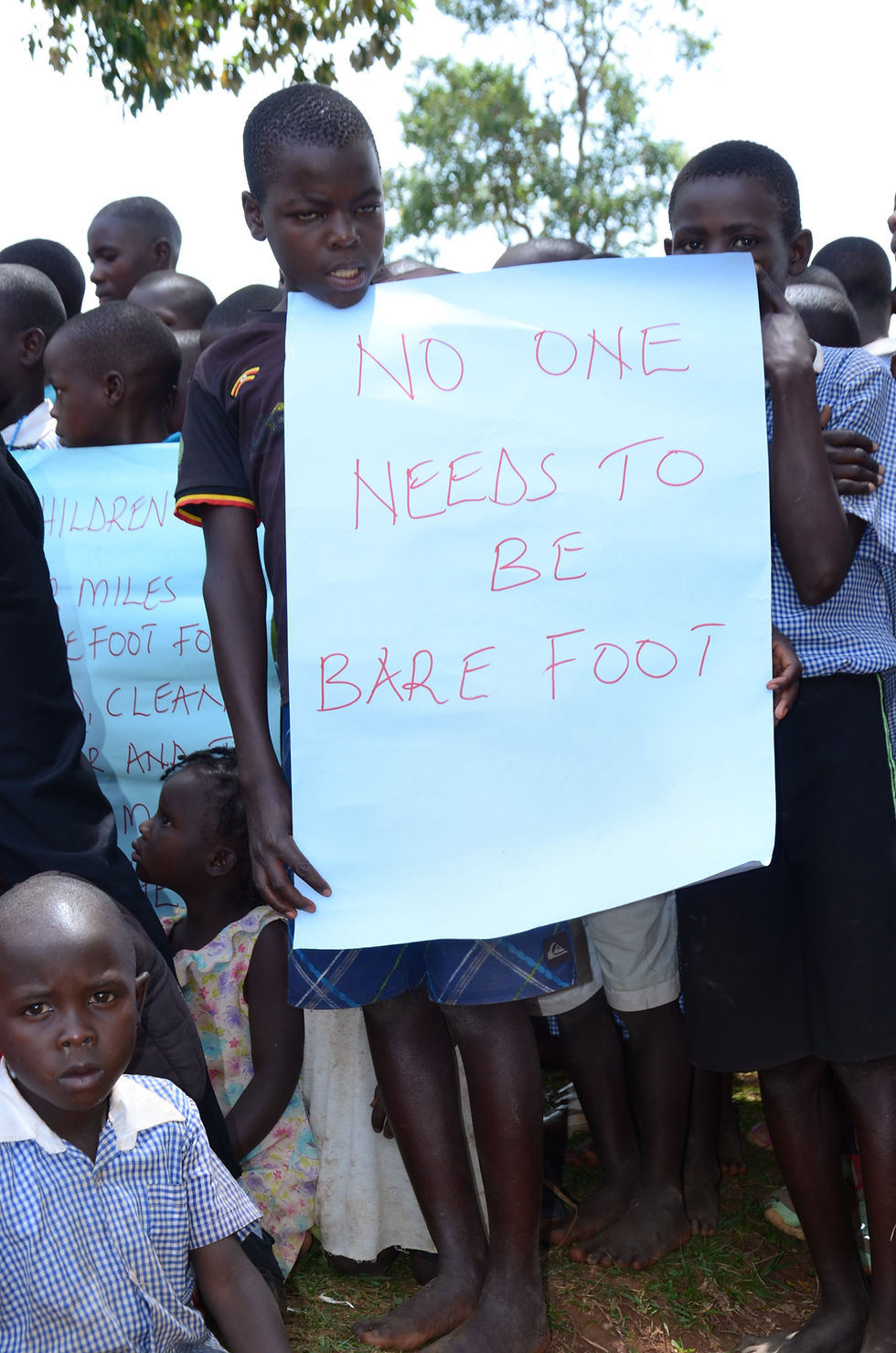 Ugandan children holding a handmade sign that says ‘No one needs to be barefoot,’ advocating for CHOCU’s shoe donation program.