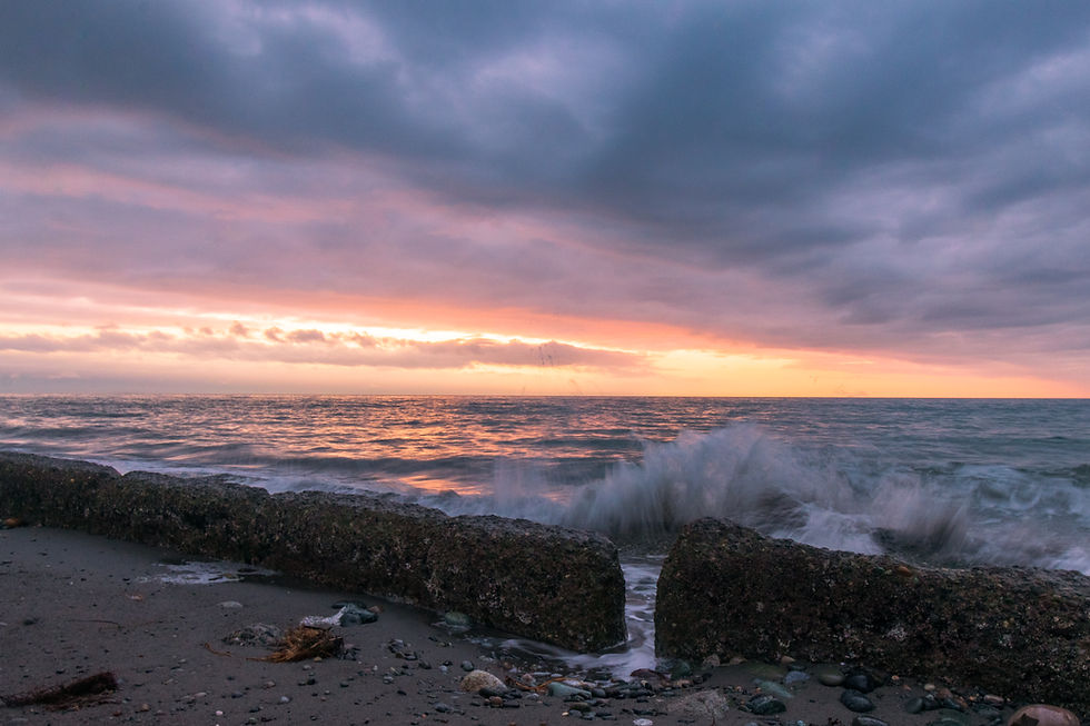 Landscape photo of a rocky beach shoreline at sunset with waves hitting a low concrete seawall, soft pastel sky with clouds