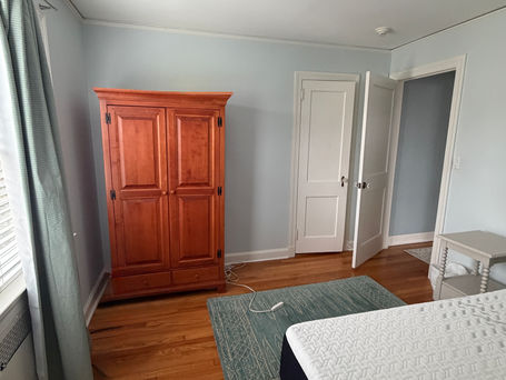 Existing guest room with items being kept - wardrobe, upholstered bed, nightstand and ceiling light and some items - rug, curtains and rods being donated.