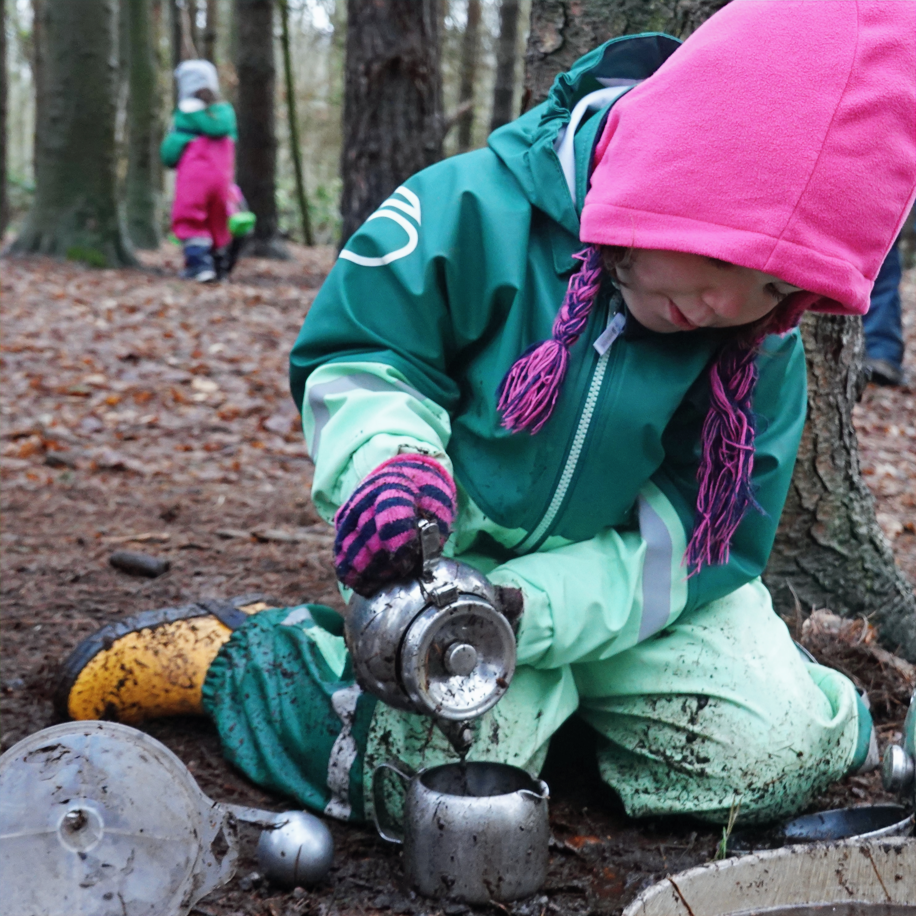 Girl in a winter hat and waterproofs pouring muddy water in the mud kitchen during outdoor preschool sensory play.