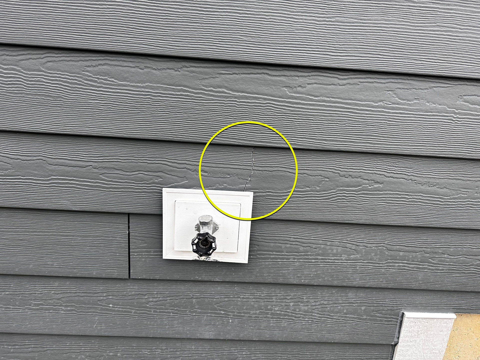 Grey horizontal siding on a home with a visible vertical crack near a water spigot, indicating potential moisture damage.