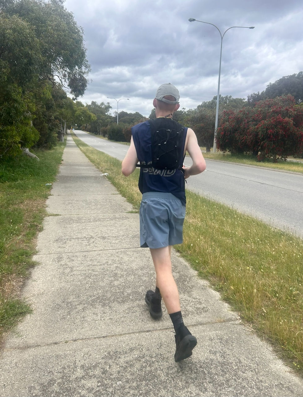 Runner seen from behind on a cloudy suburban sidewalk beside a road, wearing a cap, vest, blue shorts, and black shoes.