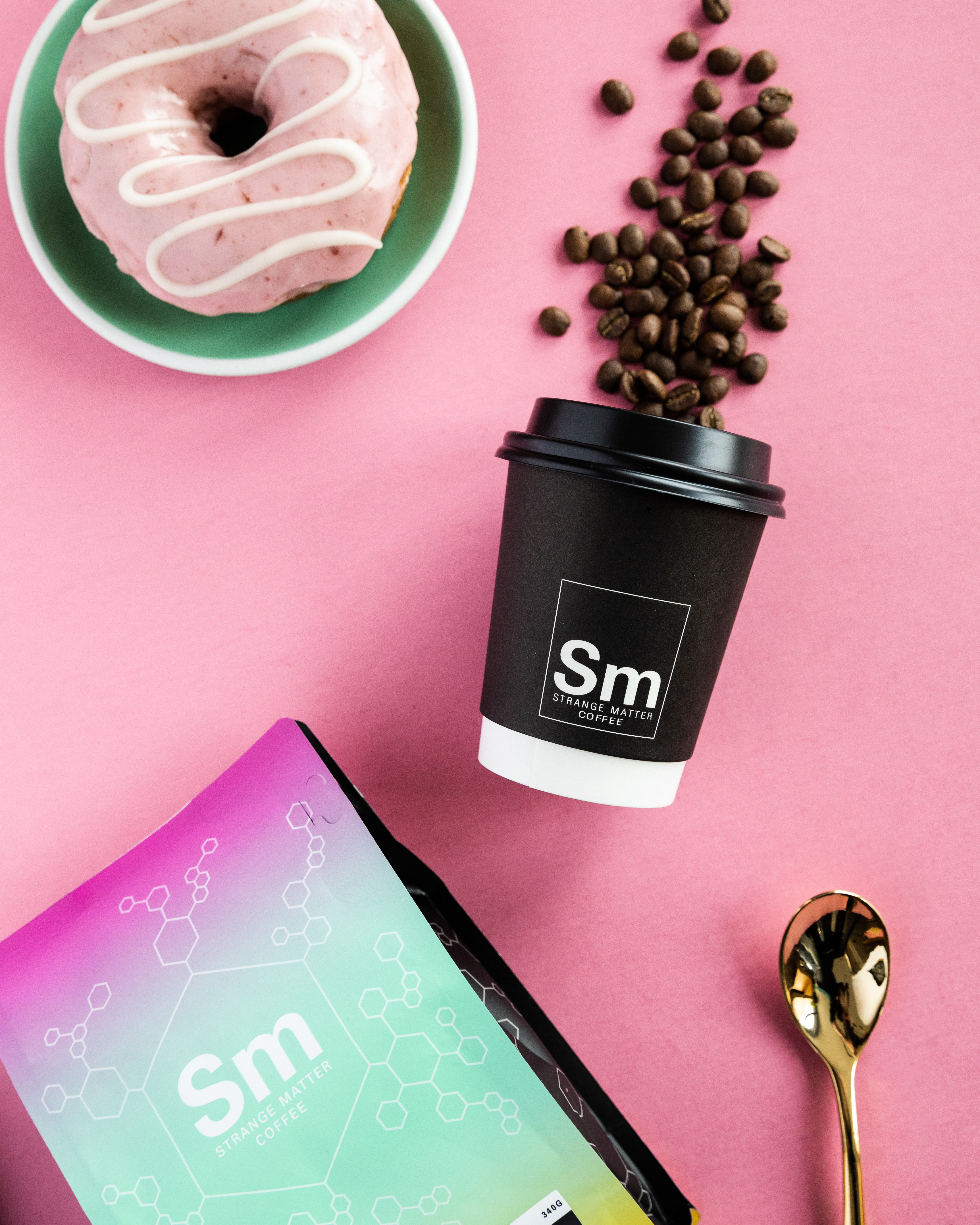 donut, coffee cup with beans, and gold spoon lay flat on pink backdrop for strange matter product photography