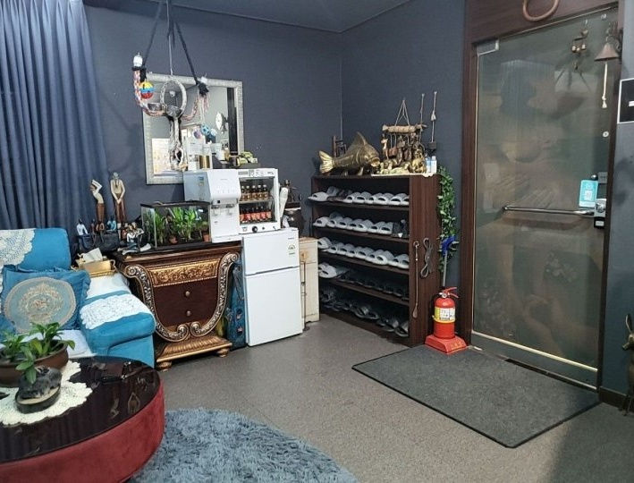 파란 소파와 장식 테이블, 신발장이 있는 실내 휴게공간. 냉장고와 유리문 옆에 소화기가 보이고 차분한 분위기.