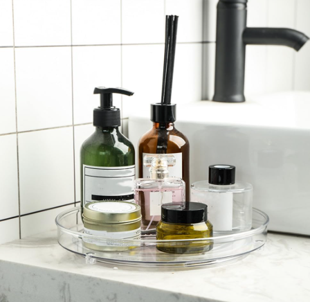 Bathroom counter with clear tray holding assorted soap and lotion bottles, jars, and pump dispensers beside a black faucet.