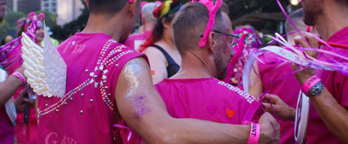 People in pink outfits with glitter, wearing bunny ears and angel wings, hug at a festive outdoor event. Vibrant atmosphere with ribbons.