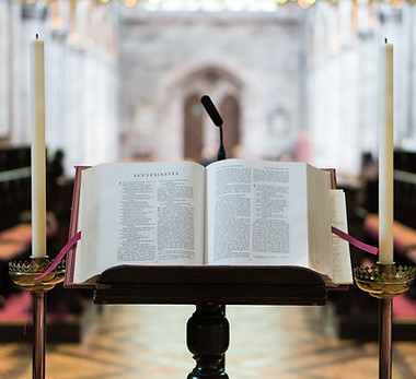 An open Bible resting on a wooden lectern, flanked by two tall lit candles inside a church sanctuary.