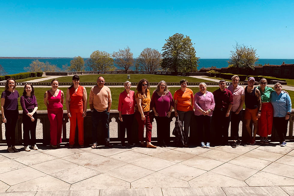 A group of people standing outdoors in a scenic location by the water, with lush greenery and a clear blue sky in the background. They are arranged in a line, smiling and dressed in a vibrant mix of colors, reflecting a sense of diversity and inclusivity. The relaxed and joyful expressions of the group capture the welcoming and supportive atmosphere of an inclusive musical community like Quorus.