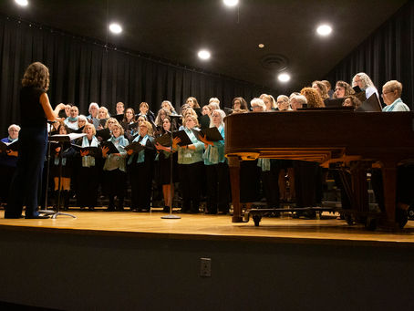 A community choir rehearses on stage under warm lights, led by a conductor with her back to the audience. Singers of all ages wear coordinated teal scarves and hold sheet music as they prepare for an upcoming concert season with Newport Sings.