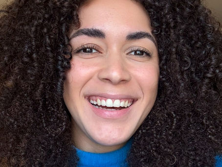 Smiling woman with curly dark hair wearing a bright blue turtleneck sweater, looking directly at the camera against a neutral background.