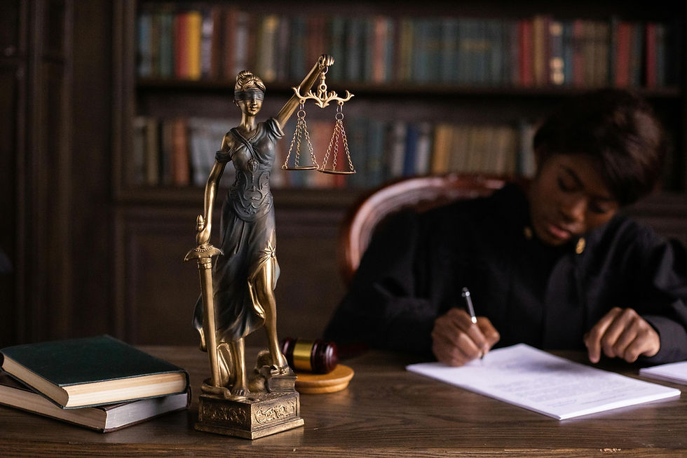 Statua della dea della giustizia con bilancia e spada su scrivania in tribunale, davanti a libri di diritto e martelletto, con giudice che scrive documento legale — simbolo di giustizia, legge e diritto.