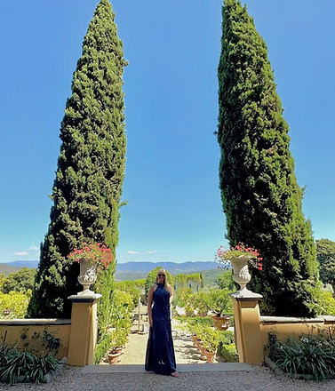 A sun-drenched view of a person in a dark dress walking through a lush Italian garden. The path is framed by tall, slender cy