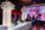 Elaborate event catering setup with a long illuminated buffet table, silver chafing dishes, tall red floral centerpieces, and a "VINTAGE VOGUE" backdrop.