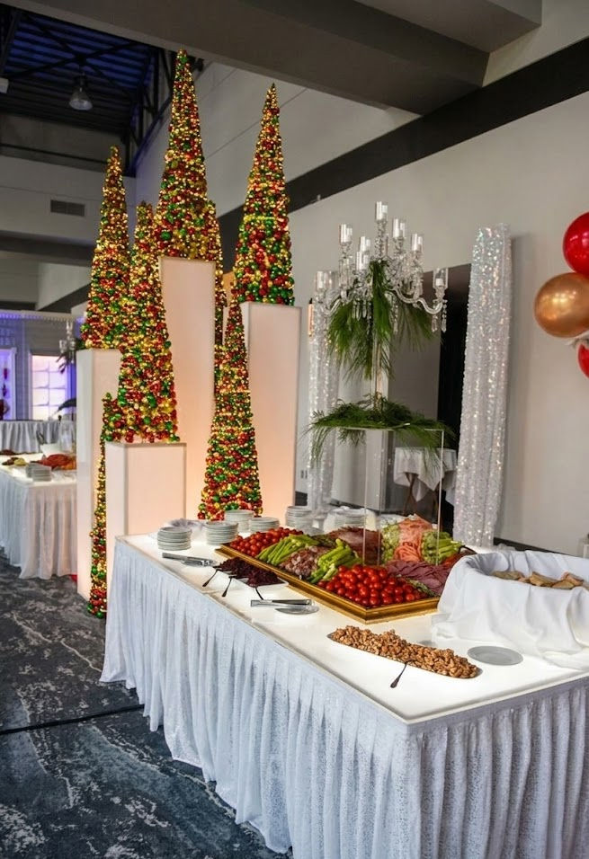 Snack table set up with a large variety of finger foods, white table cloth and a break with a couple of lit Christmas trees in between the tables.