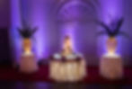 A wedding cake displayed in the middle table with two smaller tables on both sides lit with white lights in dim lighting.