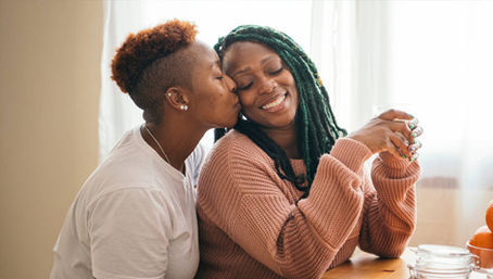 Healthy Love: one woman kisses her partner’s cheek as they smile at a sunny kitchen table, sharing a warm, supportive moment.