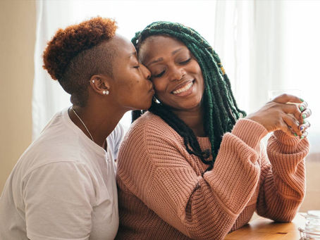 Healthy Love: one woman kisses her partner’s cheek as they smile at a sunny kitchen table, sharing a warm, supportive moment.