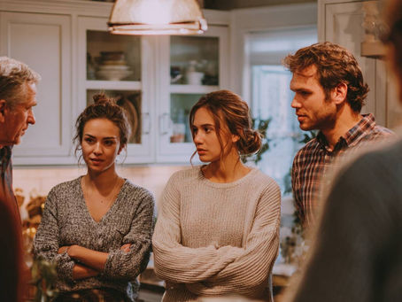 A tense family discussion taking place in a warmly lit kitchen. Several adults stand in a loose circle, with two young women in the center crossing their arms and looking upset. An older man appears to be speaking, while others listen with serious expressions. The atmosphere suggests conflict or emotional tension during a family gathering.