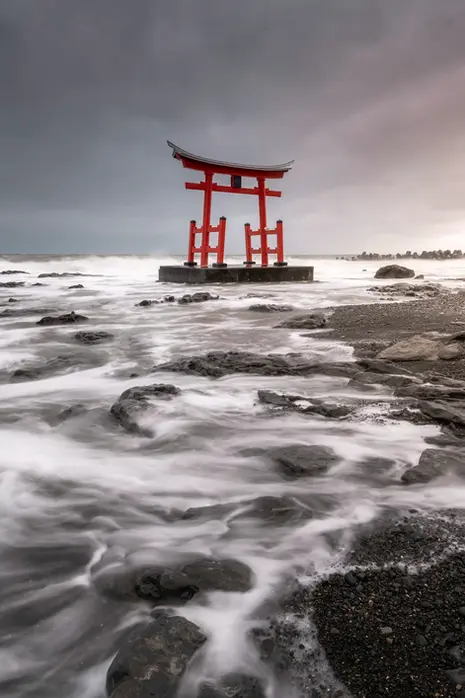 Red torii gate of Toyosaki Kompira Shrine in Hokkaido standing in ocean waves under a moody sky, long exposure seascape.