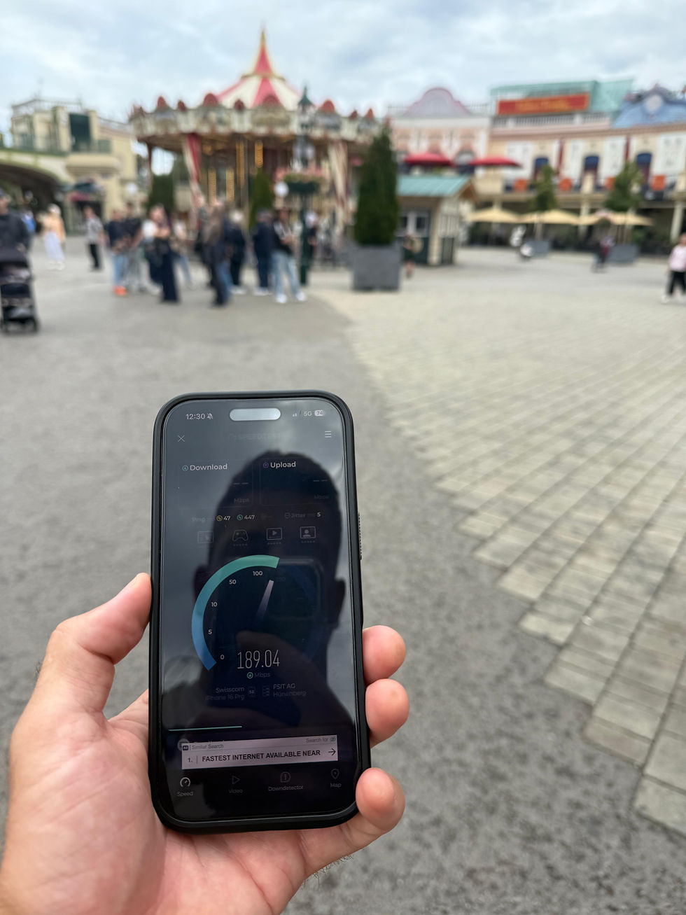 Smartphone mit laufendem Speedtest im Wiener Prater vor Fahrgeschäften und Gebäuden