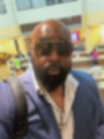 Darwin Tanksley in a blue suit jacket and white shirt wearing sunglasses, with a wooden cross necklace, standing in a bank lobby area with a confident expression.