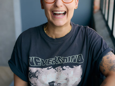 Smiling person with orange glasses and tattoos wears a Nevermind T-shirt, sitting indoors by a window.