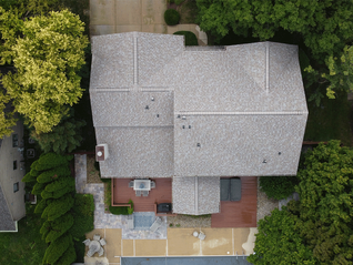 Aerial view of a house with a gray roof, surrounded by trees. Backyard features a patio with a table and chairs, and a pool area.