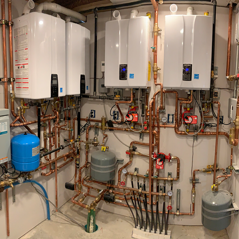 Utility room with four wall-mounted boilers, copper pipes, pumps, and tanks connected in a dense heating system.