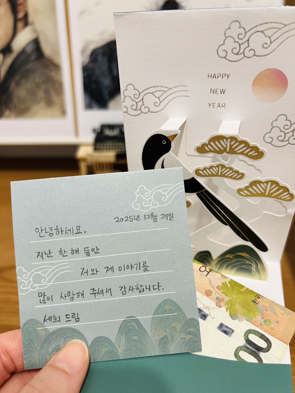 hand holding a Korean happy new year card with a Korean magpie and a handwritten message with The Moon Caught in the Pine Tree illustrations in the background