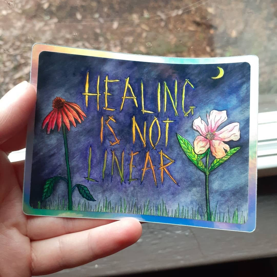 Photo of hand holding a sticker. The sticker says HEALING IS NOT LINEAR with 2 different pink flowers, a moon and grass.