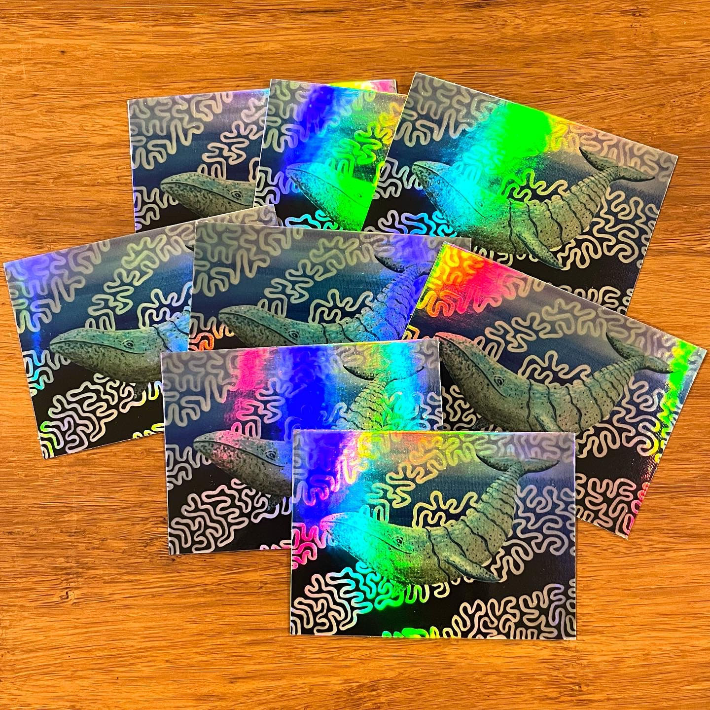 eight holographic whale stickers layered on top of each other. the holographic reflection is rainbow. the background is wood