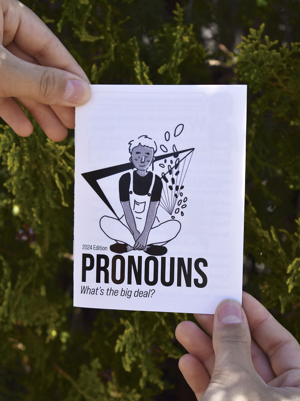 The zine held at opposite corners in front of a juniper tree. The cover has a character sitting cross-legged above the title.