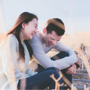 A young couple laughing together while sitting in a grassy outdoor area, dressed in sweaters.