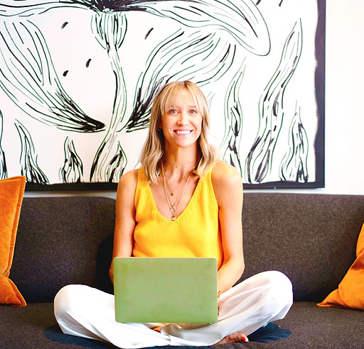 A woman sitting cross-legged with a laptop on her lap, focused on her work in a cozy indoor setting.