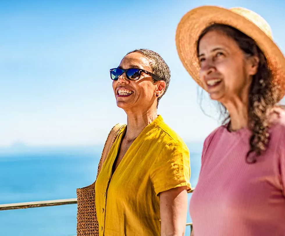 Two women smiling outdoors near the ocean. One wears sunglasses and a yellow top; the other has a hat and pink top. Bright, sunny day.