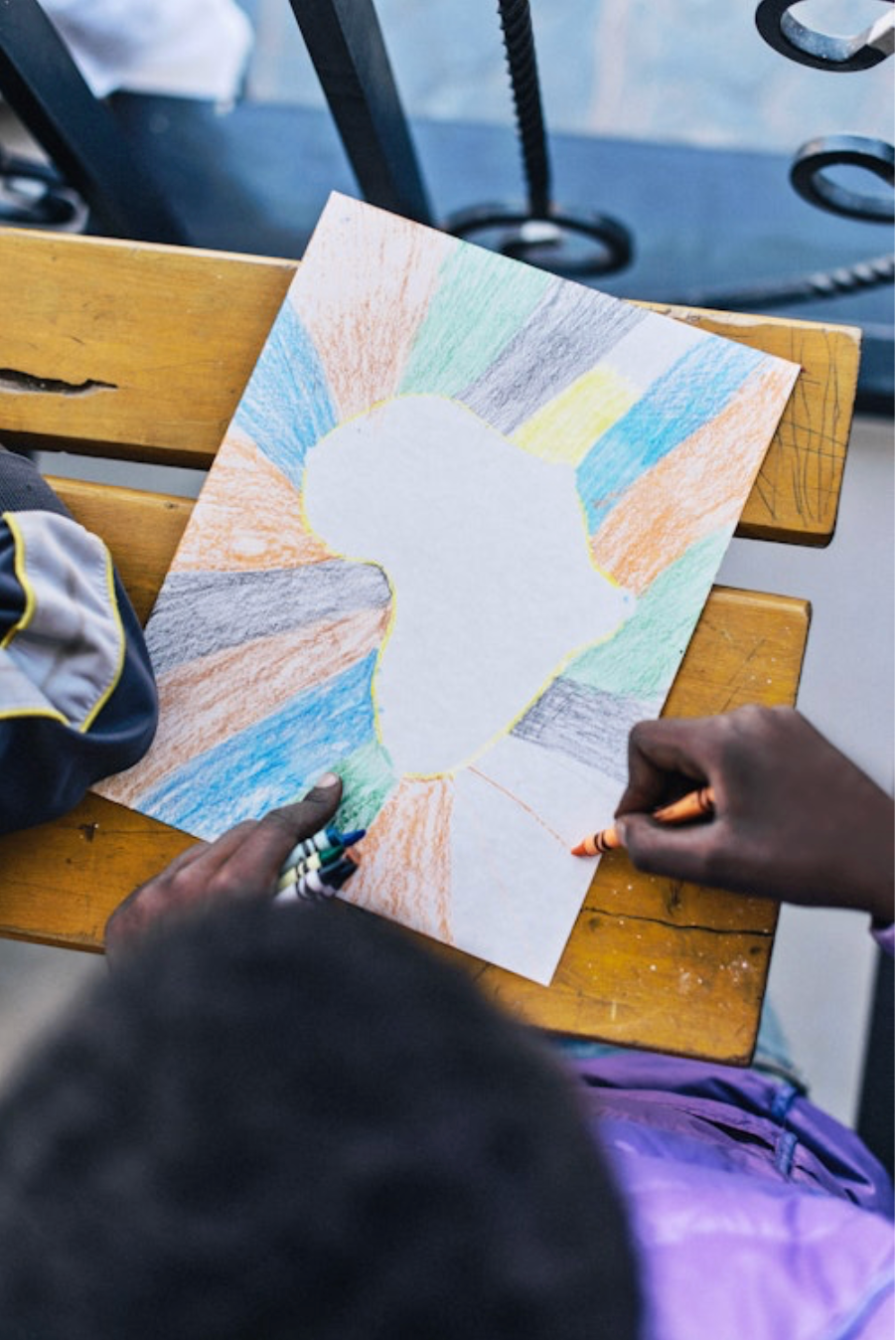 Child colors a map of Africa on paper with crayons, using bright colors, on a wooden desk. Background shows iron railing.