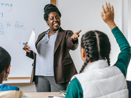 Teacher smiling and gesturing to student with raised hand in classroom. Whiteboard with blue writing in background. Engaged learning setting.