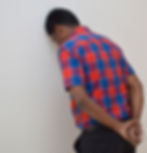 Man in red and blue plaid shirt leans forehead against a white wall, hands behind back, conveying stress. Neutral background.