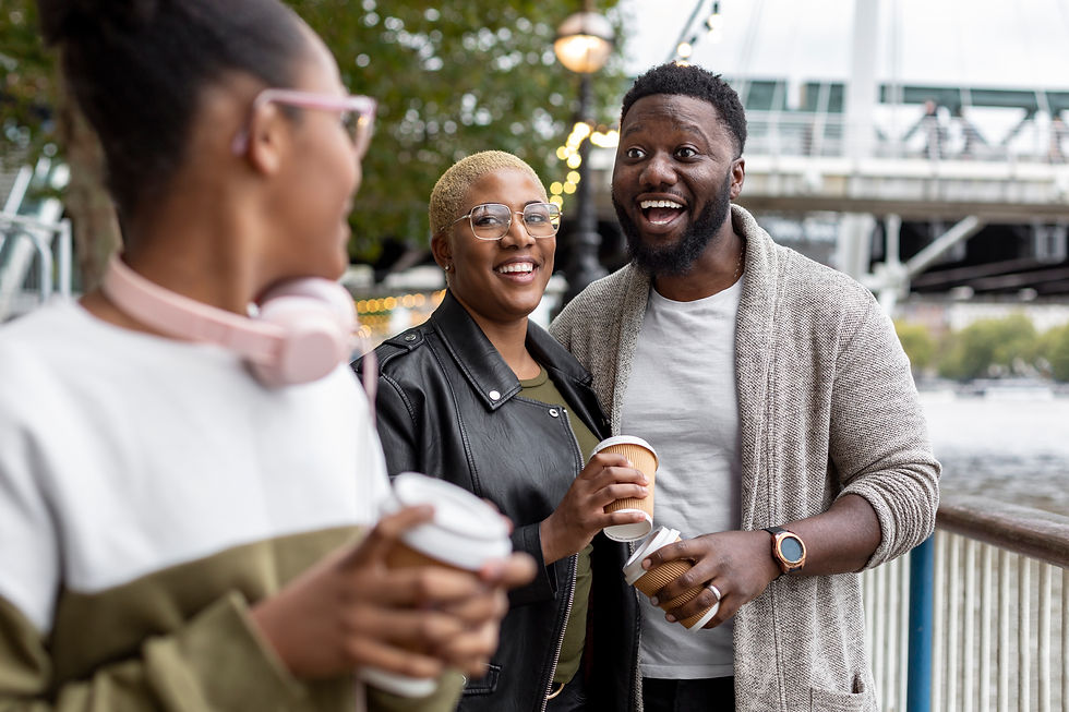Three friends chat and smile outdoors by the water, each holding coffee cups, with bridge lights and trees in the background.