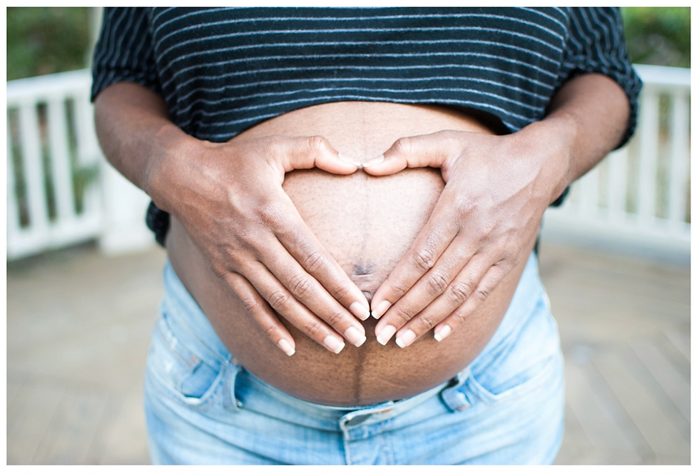 Pregnant person wearing jeans and a striped top forms a heart over their belly with hands. Wooden porch background, serene mood.