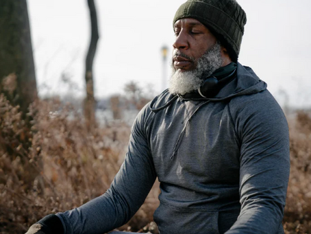 Man meditating outdoors in gray hoodie and beanie, wearing headphones. Serene expression, brown foliage and bare trees in background.