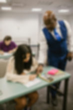 Teacher assists a student writing at a desk in a classroom. Books, a calculator, and bright pink notepad are on the desk. Another student sits behind.