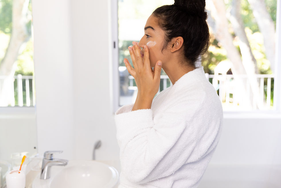 A biracial woman in a white robe is applying facial cream in a bright, sunlit white room, emphasizing the importance of establishing a consistent summer skincare routine for maintaining healthy skin