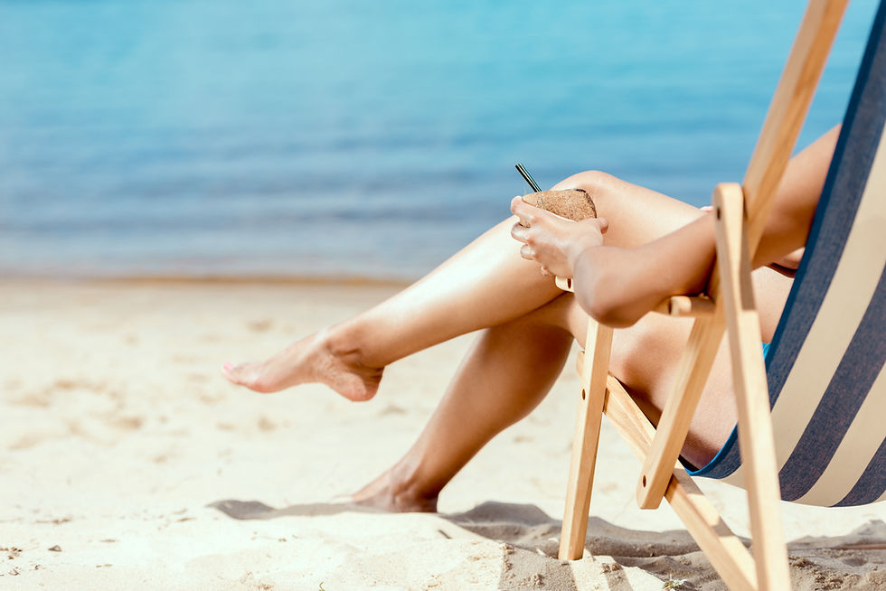 A cropped image of a woman's legs and hand holding a cocktail in a coconut shell, laying on a deck chair on a sandy beach, representing the relaxation and advanced skincare treatments available at Revital Med Spa to enhance your summer glow.