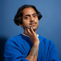 A picture of Krishna Istha, with short black hair wearing a large blue jumper in front of a blue backdrop