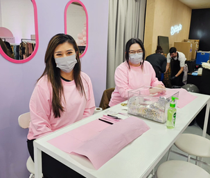 Two nail technicians in pink uniforms seated behind prepared mobile manicure tables.