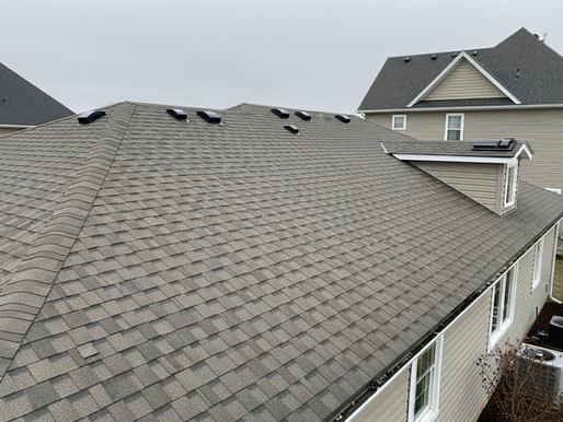 Gray shingled roof with multiple vents, under cloudy sky. Beige house in background, windows and air conditioning units visible.